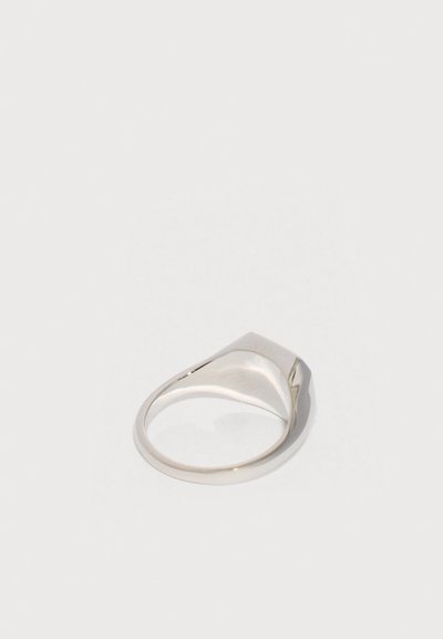 Simított, polírozott ezüst pecsétgyűrű lapos, enyhén dőlt felső felülettel, egy egyszerű világosszürke háttéren pihenve.