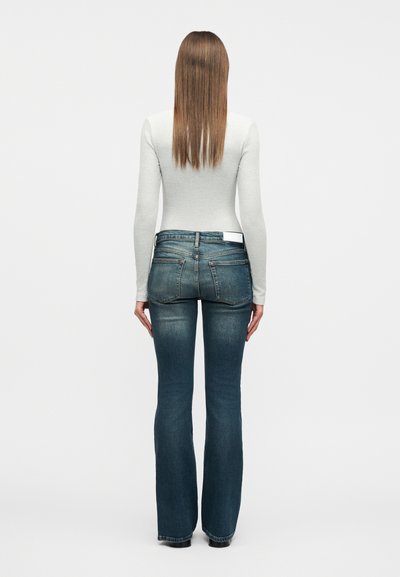 Γυναίκα με ίσια καστανά μαλλιά, φορώντας εφαρμοστό ανοιχτό γκρι μακρυμάνικο τοπ και καμπάνα μπλε τζιν, στέκεται μπροστά σε απλό λευκό φόντο.