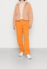 Light brown fleece jacket with a beige hooded sweatshirt and orange sweatpants. White Nike logos on the jacket and pants. White sneakers.