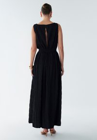 Schwarzes Maxikleid mit strukturiertem Karomuster, rundem Ausschnitt und einem Schlitz am Rücken. Elastische Taille und ausgestellter Saum. Elfenbeinfarbene Schuhe.