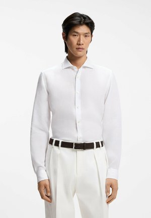 Homme portant une chemise blanche boutonnée rentrée dans un pantalon blanc plissé avec une ceinture noire, sur fond blanc uni.