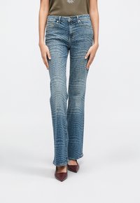 Mujer que lleva jeans de mezclilla azul con un patrón de texto "JOOP! JEANS" en todo el tejido, combinados con tacones puntiagudos burdeos y una blusa verde oliva.