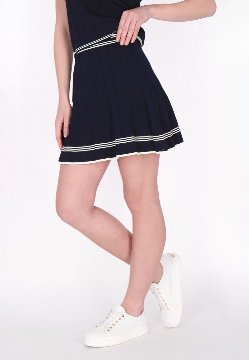 Marineblauwe geplooide jurk met een witte gestreepte tailleband, gecombineerd met witte sneakers. Glad materiaal, tot boven de knie lengte, en een uitlopende silhouet.
