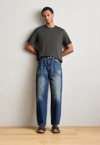 T-shirt in cotone verde scuro, vestibilità rilassata; jeans in denim azzurro chiaro, gamba dritta con design sbiadito; cintura nera; sandali scuri; sfondo in legno.