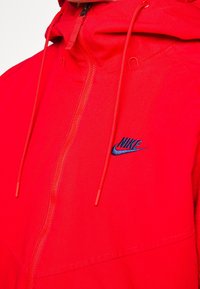 Röd Nike zip-up hoodie med dragsnören och broderad svart Nike-logotyp på bröstet, närbild.
