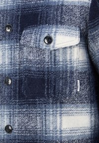 Camicia di flanella a quadri blu navy e bianca, con bottoni neri, tasca sul petto e etichetta del marchio tessuta. Tessuto testurizzato e caldo.