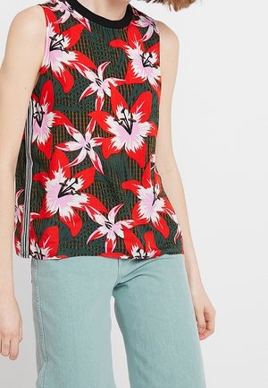 Hihaton toppi, jossa rohkeat punaiset ja vaaleanpunaiset liljat vihreällä taustalla, musta kaulus, päällä vaaleansiniset farkut seisovalla henkilöllä.