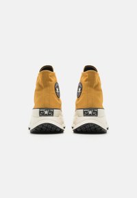 Yellow high-top sneakers with rubber soles, featuring black logo patches, textured fabric, and white accents around the sole.