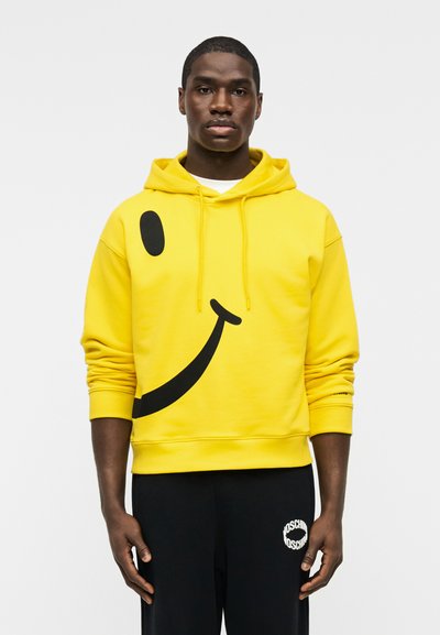 Άνδρας που φοράει κίτρινο φούτερ με κουκούλα με μεγάλο μαύρο σχέδιο χαμογελαστού προσώπου, συνοδευόμενο από μαύρο παντελόνι με λευκό λογότυπο στο αριστερό μπούτι.