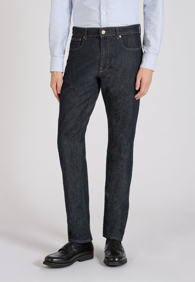 Jeans en denim bleu foncé avec une coupe droite, dotés de coutures orange et d'une fermeture à bouton à l'avant. Associés à des mocassins noirs.