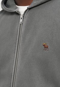 Grauer Zip-Hoodie mit weicher Textur, Kängurutasche und einem kleinen, gestickten Elchdesign auf der linken Brust.