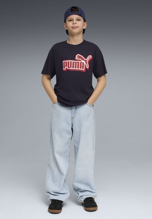 Chłopiec w granatowej koszulce Puma, jasnoniebieskich luźnych dżinsach, czarnych tenisówkach i odwróconej granatowej czapce stoi z rękami w kieszeniach.