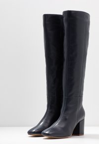 Bottes noires en cuir montant jusqu'au genou, avec un bout carré et un talon épais, présentant une texture lisse et des détails de couture minimaux.