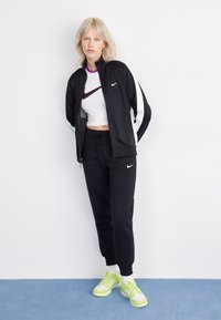 Zwarte zip-up jas met witte zijstrepen, zwarte joggingsbroek en neon groene sneakers. Korte witte top met een paars accentontwerp.