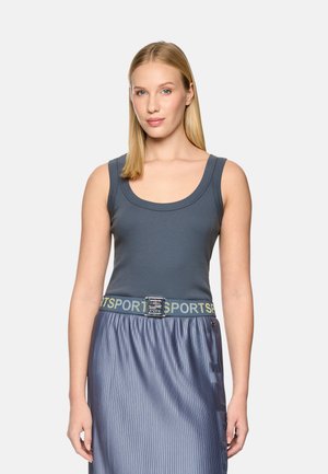 Blonde woman wearing a sleeveless gray tank top and a pleated blue skirt with a sporty belt, standing against a plain background.