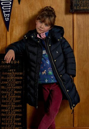 Mladé dievča s hnedými vlasmi sa opiera o drevenú stenu, má na sebe čiernu vatovanú bundu, farebnú košeľu so zvieracím motívom a červené nohavice.