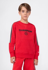 Chłopiec z krótkimi brązowymi włosami, ubrany w czerwony sweatshirt Champion i dopasowane spodnie z czarno-białymi paskami na rękawach i bokach.