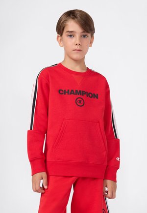 Junge mit kurzem braunem Haar, der einen roten Champion-Pullover und passende Hosen mit schwarz-weißen Streifen an den Ärmeln und Seiten trägt.