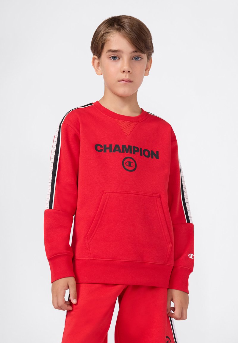 Chłopiec z krótkimi brązowymi włosami, ubrany w czerwony sweatshirt Champion i dopasowane spodnie z czarno-białymi paskami na rękawach i bokach.