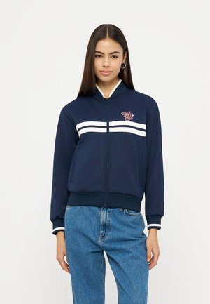 Młoda kobieta ubrana w granatową kurtkę na zamek z białymi paskami i logo, w zestawieniu z niebieskimi dżinsami z wysokim stanem, stoi na białym tle.