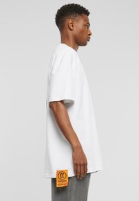Balta per didelį medvilninį T-shirt su trumpomis rankovėmis, su ryškiu oranžiniu prekės ženklo žymeklį šone. Paprastas, tiesios kirpimo dizainas.