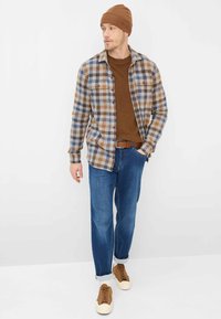 Camicia a quadri in blu, marrone e crema sopra un maglione marrone; jeans blu; scarpe marroni con suole crema; berretto marrone.