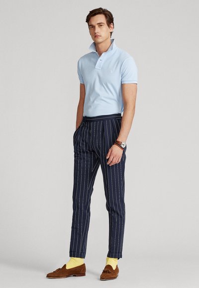 Mand iført lyseblå polo, marineblå bukser med tynde striber, brune loafers, gule sokker og et armbåndsur, står med hænderne i lommerne.