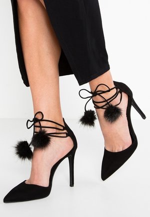Black high-heeled shoes with a pointed toe, suede material, ankle ties, and two fluffy black pom-poms on the sides.