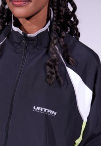 Casaco preto com fecho de correr e acentos brancos e verdes, gola alta e logótipo "URTRN" no peito. Leve, com textura suave.