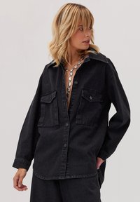 Czarna kurtka dżinsowa z kołnierzykiem, zapięciem na guziki i dwoma przednimi kieszeniami. Charakteryzuje się luźnym krojem i wyraźnie zarysowanymi ramionami.