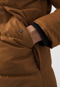 Brown puffer jacket, featuring a smooth texture, with a snap button detail near a pocket and a black ribbed cuff.