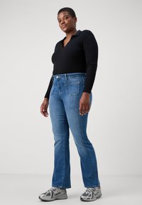 Tumman pitkähihaisen polo-paidan parina vaaleansiniset leveät farkut, joissa on takataskut ja hopeiset yksityiskohdat, ja asusteenaan harmaat urheilukengät.