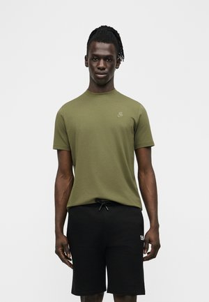 Mladý muž s tmavou pletí a dredy, oblečený v olive zelené krátké košili a černých šortkách se stahovací šňůrkou, stojící proti jednobarevnému pozadí.