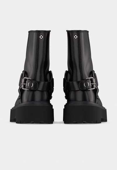 Μαύρα δερμάτινα μποτάκια αστραγάλου με χοντρές σόλες, ασημί πόρπες και επάνω οπές, εμφανιζόμενα από πίσω σε φωτεινό φόντο.