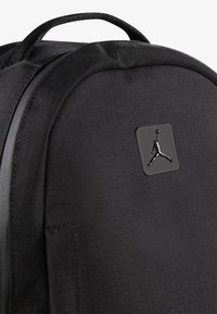 Crni ruksak od teksturirane tkanine s istaknutim crnim Jumpman logotipom na kvadratnoj oznaci. Uključuje gornju dršku za nošenje.