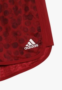 Punaiset urheilushortsit, joissa on pyöreä kuvio, ominaisuutenaan teksturoitu vyötärö ja valkoinen Adidas-logo vasemmassa alareunassa.