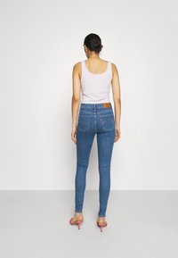 Top de tirantes lila claro con escote en forma de olas, combinado con jeans ajustados azules que tienen bolsillos traseros y un parche marrón. Zapatos de tacón rosas.