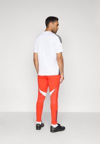 Conjunto deportivo que incluye una camiseta blanca de manga corta con franjas negras laterales y pantalones ajustados rojos con detalles triangulares blancos y cremalleras laterales.