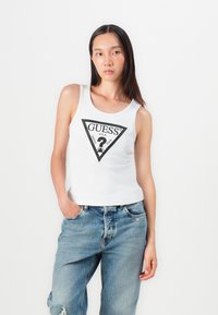 Top alb, cu dungi, fără mâneci, cu un logo triunghiular negru care conține text. Asortat cu blugi din denim ușor albastri, deteriorați, având un croi lejer.