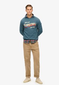 Sudadera azul con gráfico de "Ojalá estuvieras aquí", superpuesta a una camisa de cuadros. Pantalones beige y zapatillas crema completan el look casual.