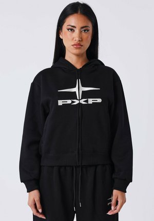 Een vrouw staat rechtop met haar gezicht naar voren, gekleed in een zwarte ritshoodie en bijpassende broek met een zilveren "PXP"-logo op de borst en boven de dij.