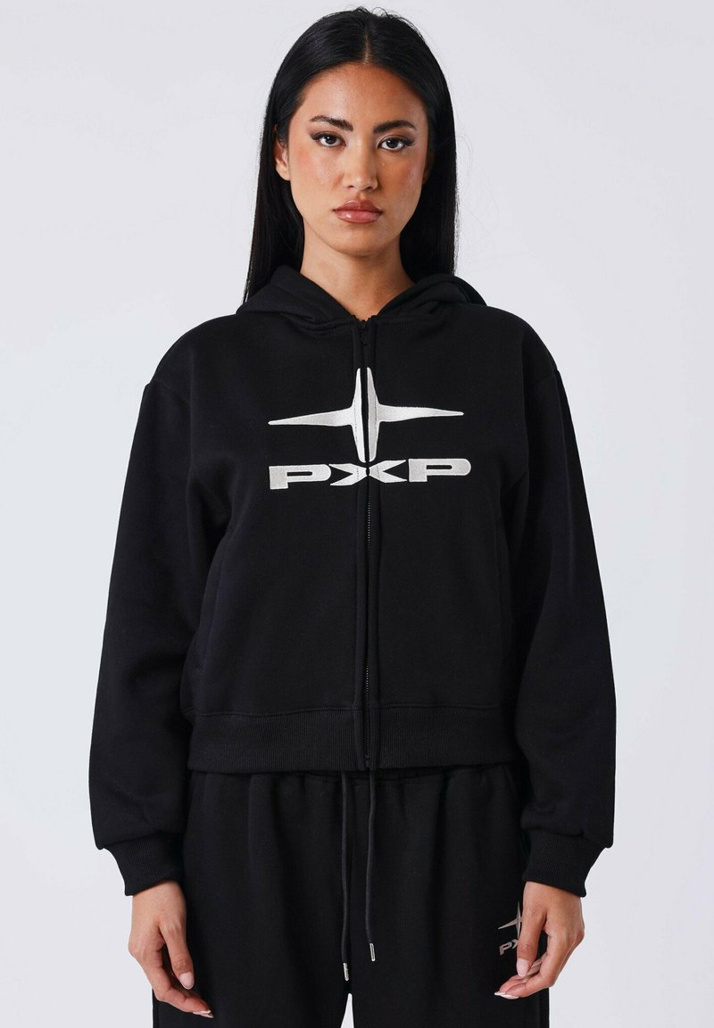 Femme se tenant face à l'avant, portant un sweat à capuche zippé noir et un pantalon assorti avec le logo argenté "PXP" sur la poitrine et la cuisse.