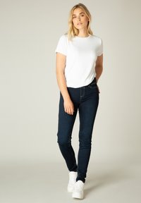 Witte T-shirt met korte mouwen, gecombineerd met donkerblauwe skinny jeans. Het model draagt witte sneakers en staat tegen een effen achtergrond.