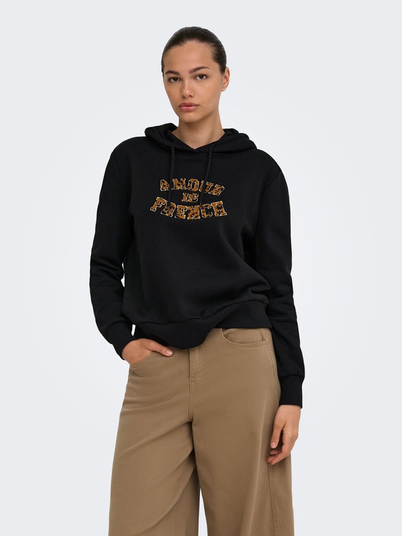 Femme portant un sweat à capuche noir avec le texte "AMOUR IS FRENCH" et un pantalon large beige, debout avec une main dans la poche devant un fond uni.
