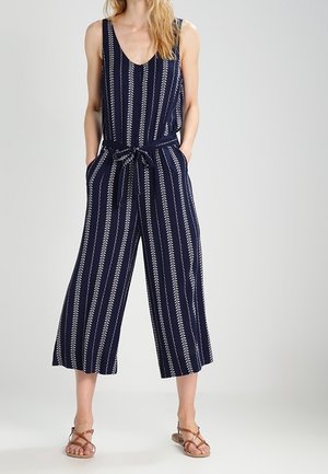 Kvinde iført ærmeløs marineblå jumpsuit med hvide lodrette bladmønstre, bindebånd i taljen, brede korte ben og brune sandaler med stropper.