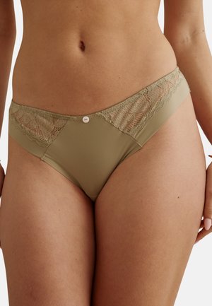 Femme portant un ensemble de sous-vêtements style bikini en dentelle beige et tissu lisse, debout avec les mains détendues sur les côtés.