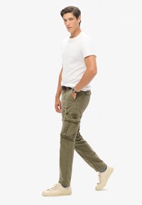 Pantalones cargo de tela verde oliva con bolsillos y detalles de costura. Combinados con una camiseta ajustada blanca y zapatillas de deporte en un tono beige claro.