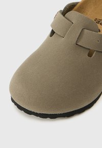 Scarpa slip-on in suede taupe con punta chiusa, caratterizzata da una sola fascia e una superficie testurizzata. Suola in gomma nera con una leggera curvatura.