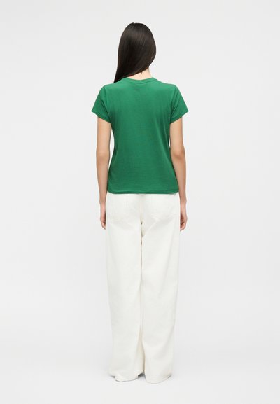 Γυναίκα με μακριά σκουρόχρωμα μαλλιά, στέκεται με την πλάτη προς τα εμπρός, φοράει πράσινο κοντομάνικο πουκάμισο και άνετο λευκό παντελόνι, μπροστά σε απλό φόντο.