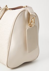Sac à main beige clair avec un motif de rose matelassé, texture en cuir lisse, accessoires en or, fermetures éclair et une étiquette de bagage amovible.